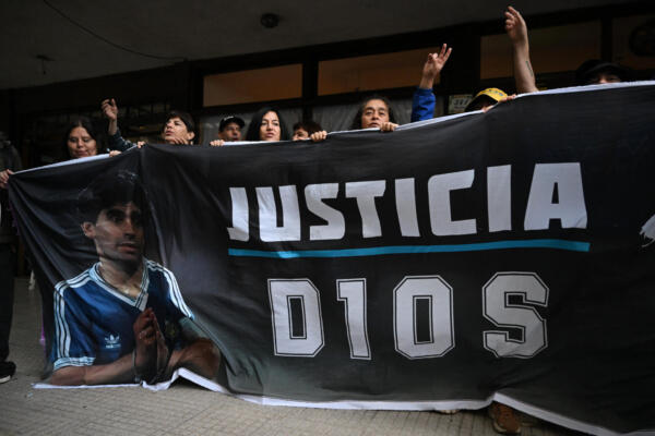 Aficionados gesticulan mientras sostienen una bandera con la imagen del fallecido Diego Maradona frente al juzgado de San Isidro, antes del juicio por la muerte de la leyenda del fútbol argentino. AFP
