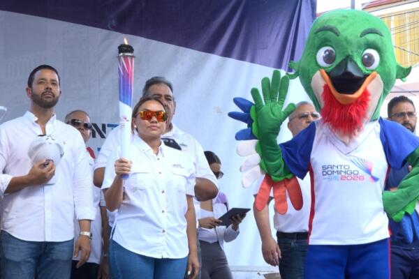 La gobernadora de Moca, Patricia Rivera, sostiene la antorcha de los Juegos Santo Domingo 2026, mientras Colí, la mascota oficial, saluda al público.

