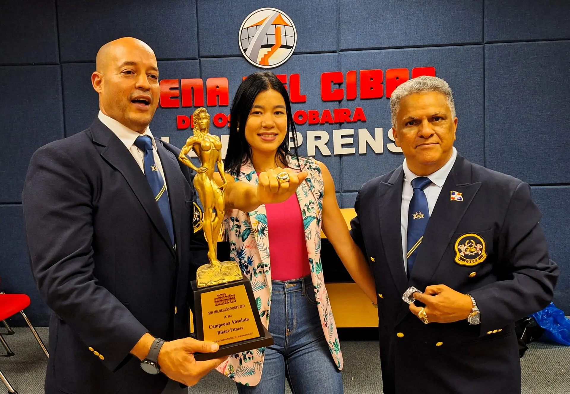 La atleta Gionmy Chol recibe el gran absoluto bikini’s XIII Mr Región Norte’2023 y el anillo correspondiente de manos de los señores, Tony Peña R, presidente de FDFF y Juan Carlos Rodríguez Ferreira, representante de IFBB Academy Caribean