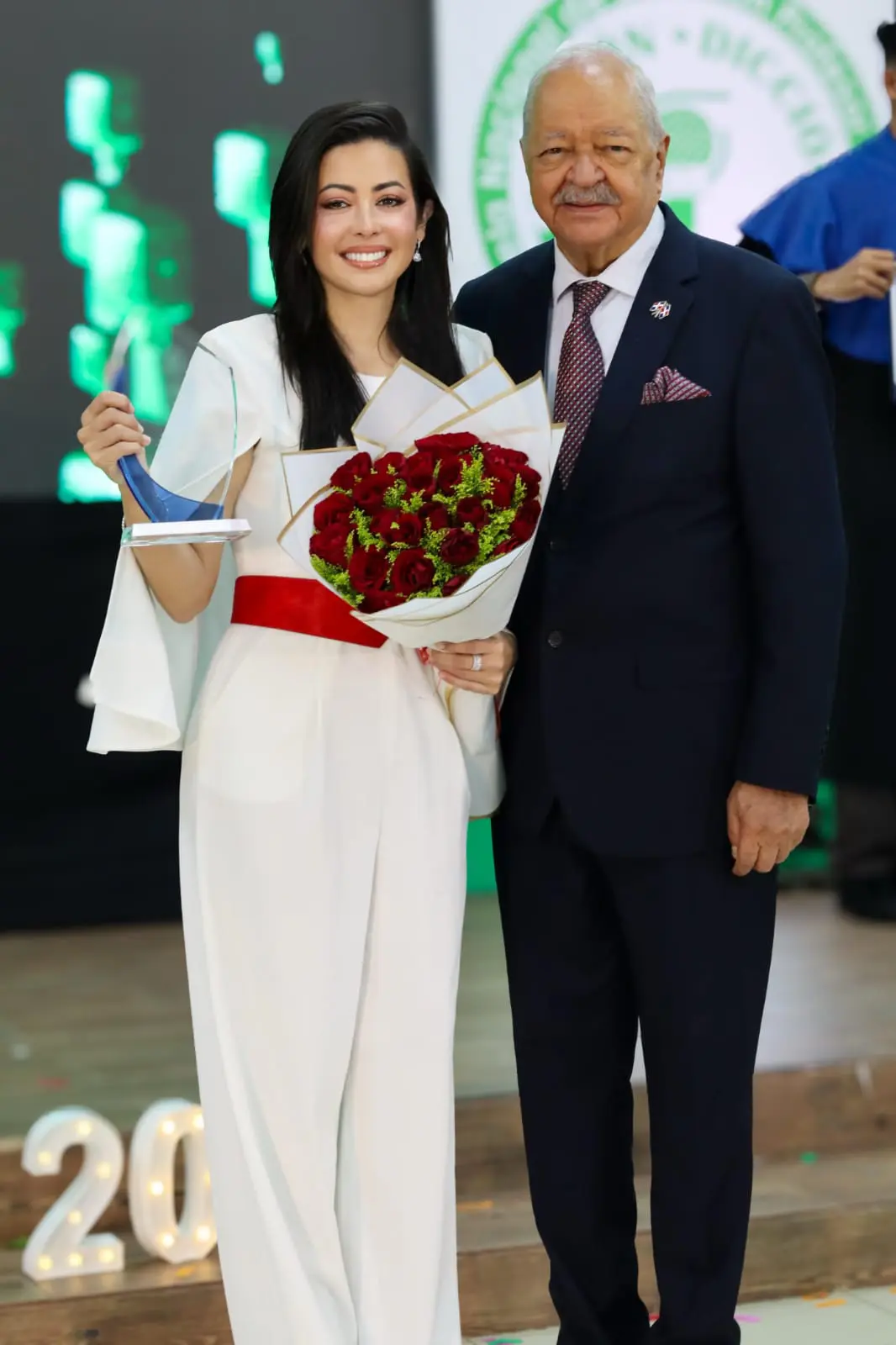 Dafne Guzmán recibe de las manos de Carlos Cepeda Suriel la placa de reconocimiento y un ramo de rosas rojas. FUENTE EXTERNA