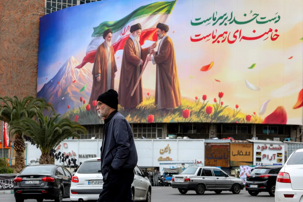 Un hombre camina cerca de una pancarta exhibida en la plaza Valiasr, en el centro de Teherán, el 10 de marzo de 2026, que representa al difunto líder supremo de Irán, el ayatolá Ruhollah Jomeini (izq.), observando cómo su sucesor, el difunto ayatolá Alí Jamenei (centro), entrega una bandera nacional a su hijo y nuevo líder supremo, Mojtaba Jamenei (der.).
